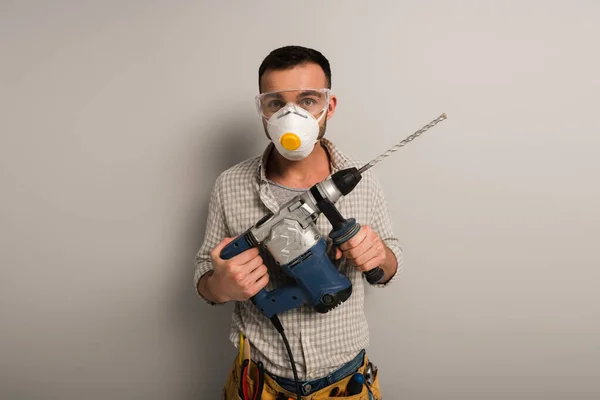 Trabajador manual emocional en la máscara de seguridad celebración de taladro eléctrico en gris - foto de stock