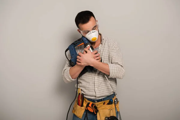 Trabalhador manual com olhos fechados em máscara de segurança abraçando broca elétrica em cinza — Fotografia de Stock