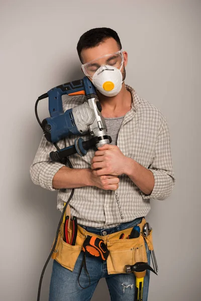 Manual worker with closed eyes in safety mask holding electric drill on grey — Stock Photo