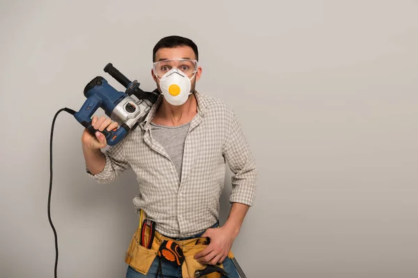 Emotional manual worker in safety mask with tool belt holding electric drill on grey — Stock Photo