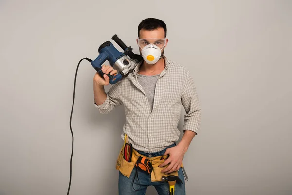 Confident manual worker in safety mask holding electric drill on grey — Stock Photo