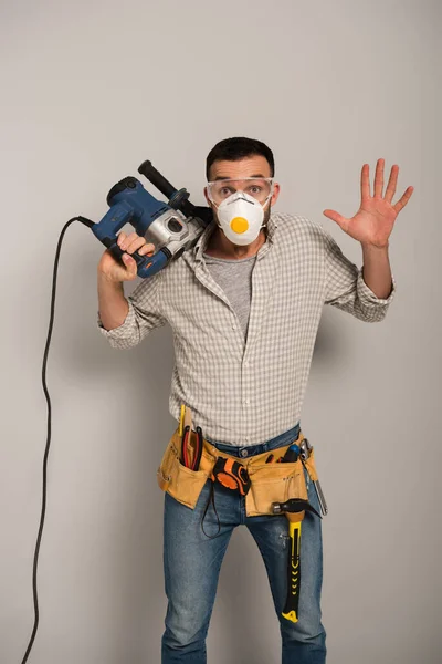 Emotionaler Arbeiter in Schutzmaske hält Elektrobohrer auf Grau — Stockfoto