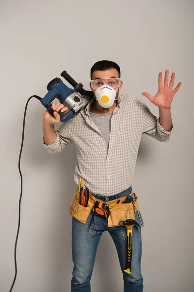 Trabajador manual emocional en la máscara de seguridad celebración de taladro eléctrico en gris - foto de stock