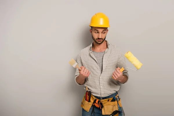 Skeptical manual worker in hardhat holding paint roller and brush on grey — Stock Photo