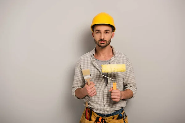 Trabajador manual masculino en hardhat sosteniendo rodillo de pintura y pincel en gris - foto de stock