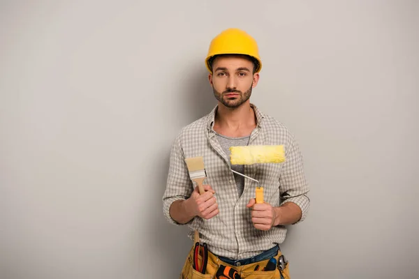 Männliche Handarbeiter in Harthut halten Farbroller und Pinsel auf grau — Stockfoto