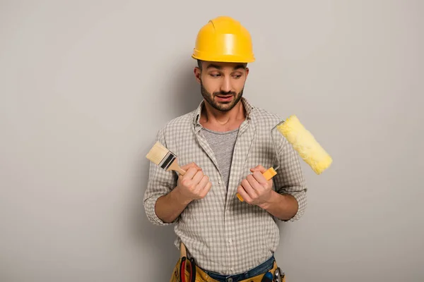 Obrero escéptico en hardhat sosteniendo rodillo de pintura y pincel en gris - foto de stock