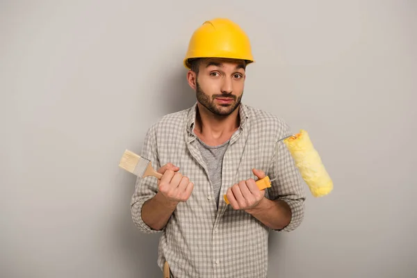 Confused workman in protective helmet holding paint roller and brush on grey — Stock Photo
