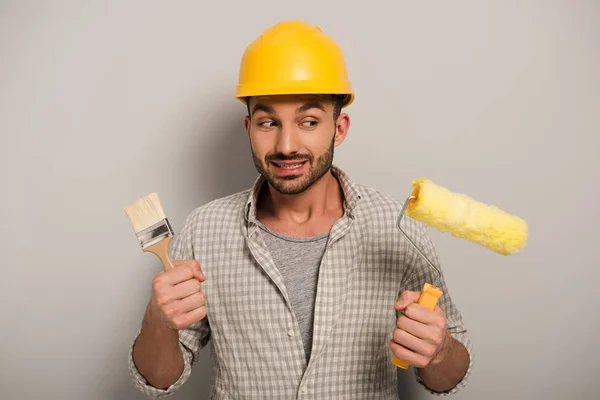 Thoughtful manual worker in hardhat looking at paint roller and brush on grey — Stock Photo
