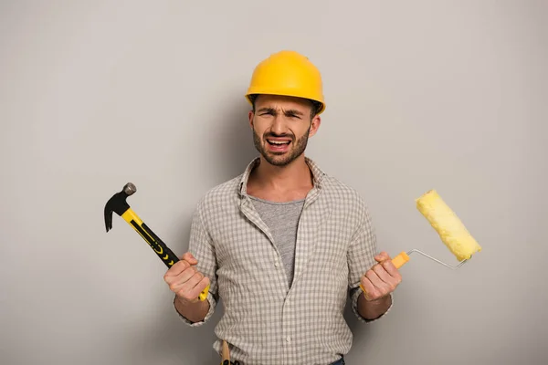 Trabajador manual estresado en hardhat sosteniendo rodillo de pintura y martillo en gris - foto de stock