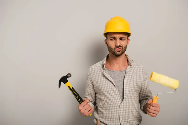 Trabalhador manual pensativo em hardhat segurando rolo de pintura e martelo em cinza — Fotografia de Stock