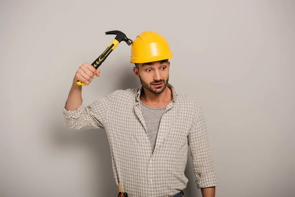 Вдумчивый ремонтник в шлеме держит молоток на сером — стоковое фото