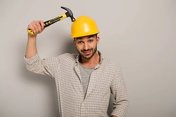Confused repairman in helmet holding hammer on grey — Stock Photo