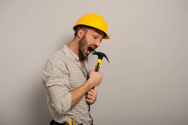 Emotional repairman in yellow helmet biting hammer on grey — Stock Photo