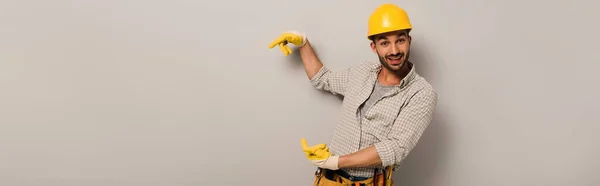 Panoramic shot of surprised manual worker in hardhat and safety gloves pointing on grey — Stock Photo