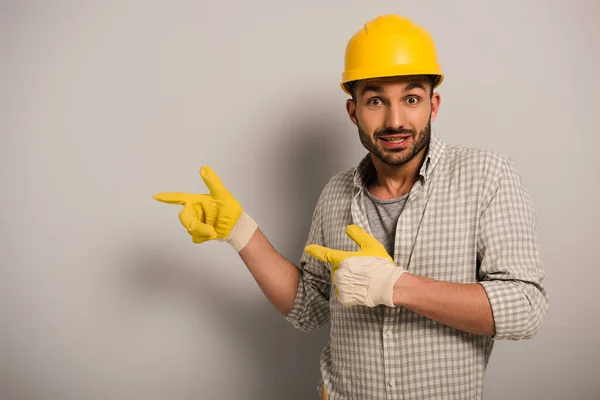 Überraschter Arbeiter mit Helm und Schutzhandschuhen zeigt auf Grau — Stockfoto