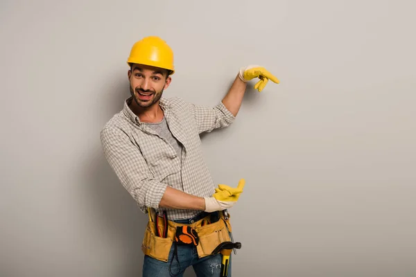 Trabalhador manual animado em chapéu duro e luvas de segurança apontando em cinza — Fotografia de Stock