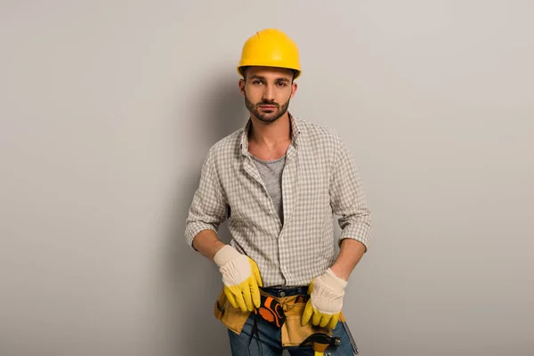 Впевнений працівник у шоломі та рукавичках з поясом інструментів на сірому — стокове фото