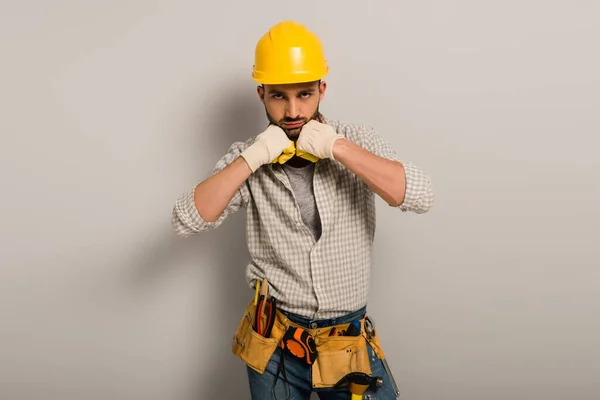 Schöner Arbeiter in Harthut und Handschuhen mit Werkzeuggürtel auf grau — Stockfoto