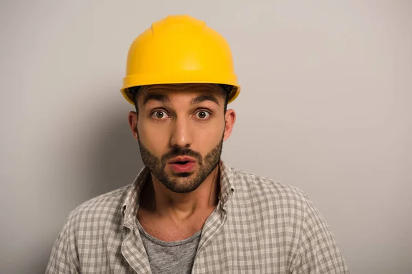 Handsome shocked manual worker in yellow hardhat on grey — Stock Photo