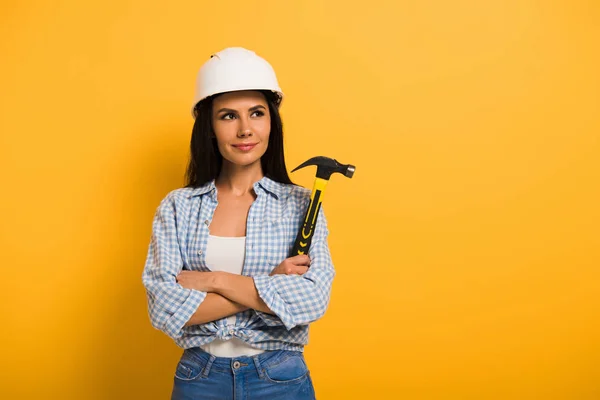 Femme de travail positive dans le casque tenant marteau avec bras croisés sur jaune — Photo de stock