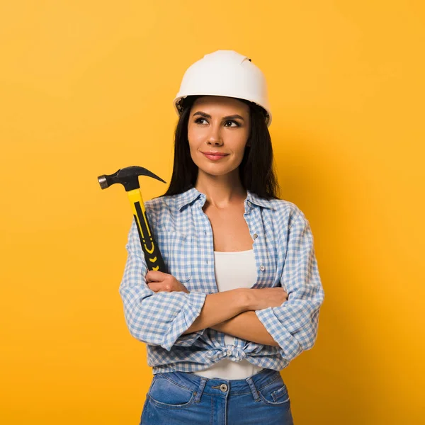 Femme de travail joyeuse dans le casque tenant marteau avec les bras croisés sur jaune — Photo de stock