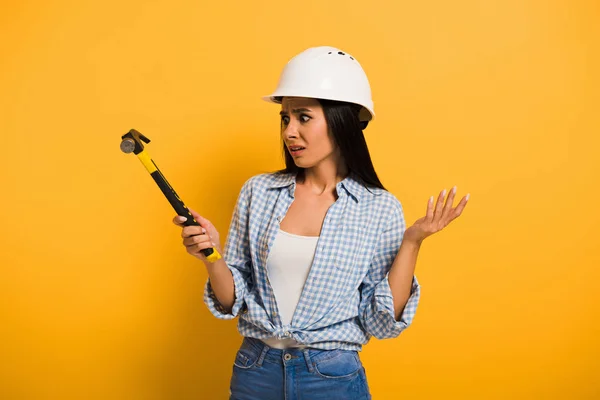 Confused female worker in helmet holding hammer on yellow — Stock Photo