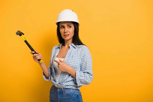 Frustrierte Arbeiterin mit Helm hält Hammer auf gelb — Stockfoto