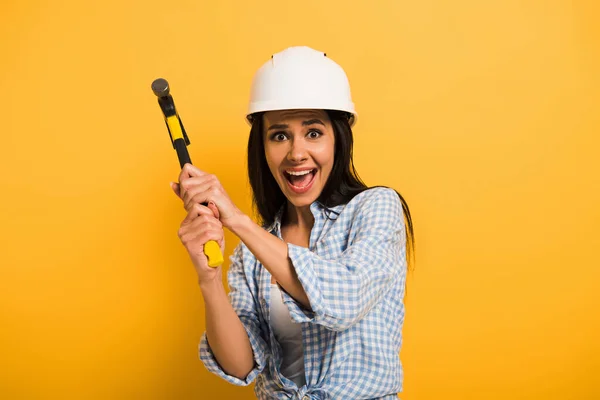 Excited female manual worker in helmet holding hammer on yellow — Stock Photo