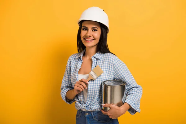 Femme de travail gaie dans le casque tenant la boîte de peinture et brosse sur jaune — Photo de stock
