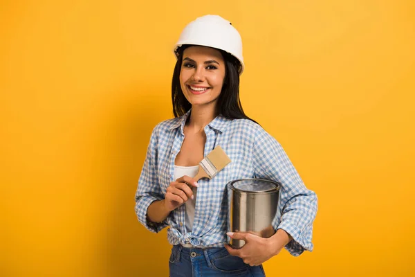 Femme de travail gaie dans le casque tenant la boîte de peinture et brosse sur jaune — Photo de stock