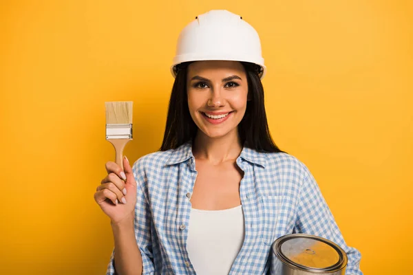 Femme de travail souriante dans le casque tenant la boîte de peinture et brosse sur jaune — Photo de stock