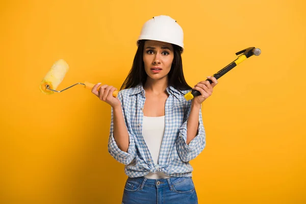 Trabalhadora frustrada no capacete segurando martelo e rolo de pintura no amarelo — Fotografia de Stock