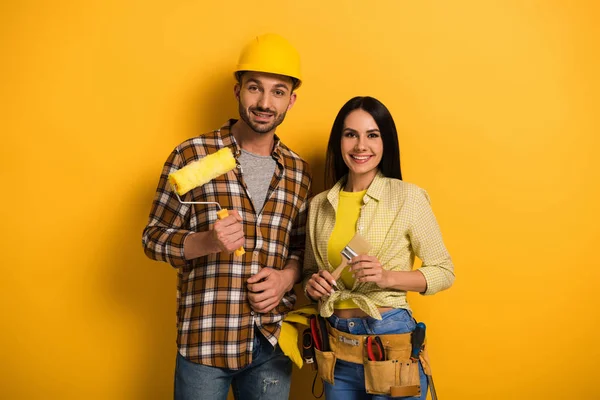 Profissionais sorrindo trabalhadores manuais segurando rolo de pintura e escova no amarelo — Fotografia de Stock