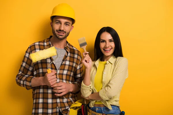 Happy manual workers holding paint roller and brush on yellow — Stock Photo