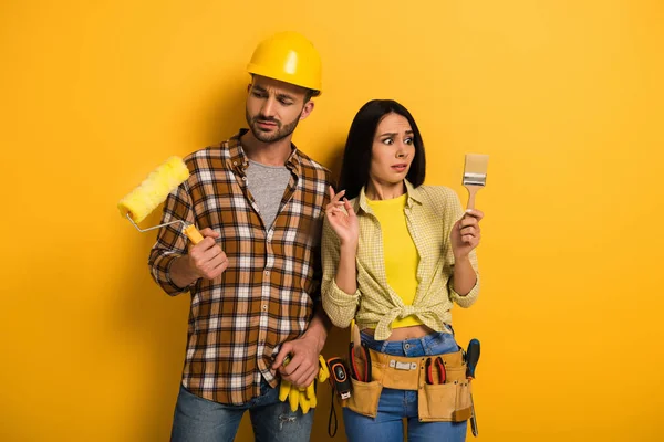 Trabajadores manuales confundidos mirando rodillo de pintura y pincel en amarillo - foto de stock