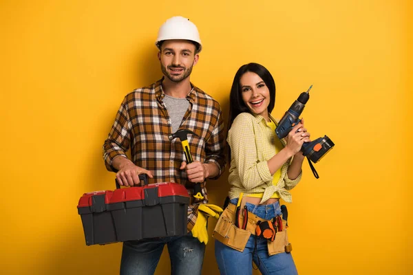 Sonrientes trabajadores manuales sosteniendo caja de herramientas y taladro eléctrico en amarillo - foto de stock