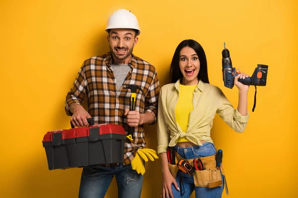 Animado handywoman e faz-tudo segurando caixa de ferramentas e broca elétrica no amarelo — Fotografia de Stock