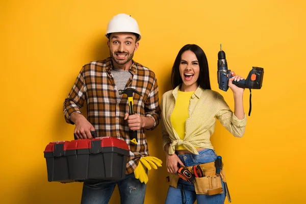 Trabajadores manuales excitados sosteniendo caja de herramientas y taladro eléctrico en amarillo - foto de stock