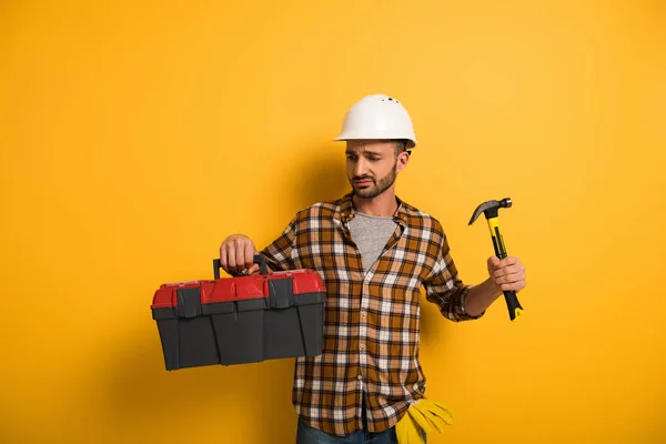 Operaio manuale frustrato in hardhat che tiene la cassetta degli attrezzi e martello su giallo — Foto stock