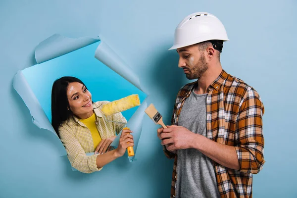 Trabalhador emocional com pincel de pintura e mulher com rolo de pintura em papel rasgado, isolado em azul — Fotografia de Stock