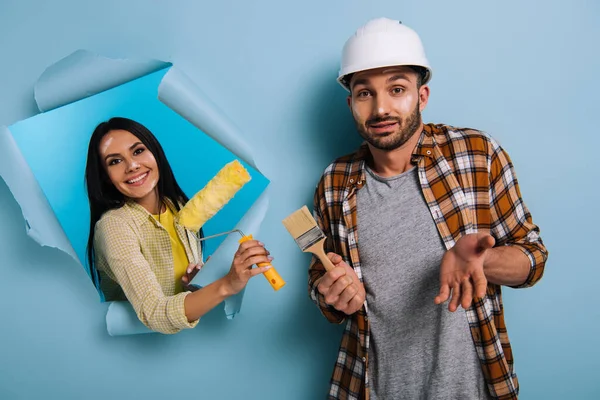 Workman with paint brush and woman with paint roller in torn paper, isolated on blue — Stock Photo