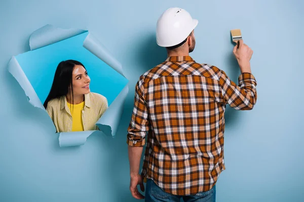 Mujer en papel desgarrado mirando al obrero con pincel, aislado en azul - foto de stock