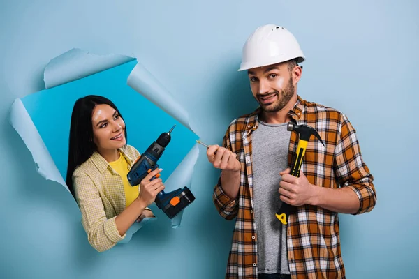 Emotional workman with hammer and woman with electric drill in torn paper, isolated on blue — Stock Photo