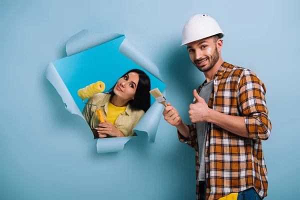 Усміхнений працівник з пензлем для фарби, що показує великий палець і жінка з фарбою в рваному папері, ізольовані на синьому — стокове фото