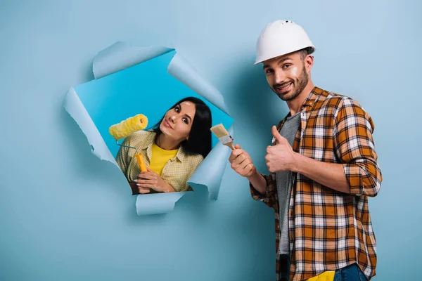 Smiling workman with paint brush showing thumb up and woman with paint roller in torn paper, isolated on blue — Stock Photo
