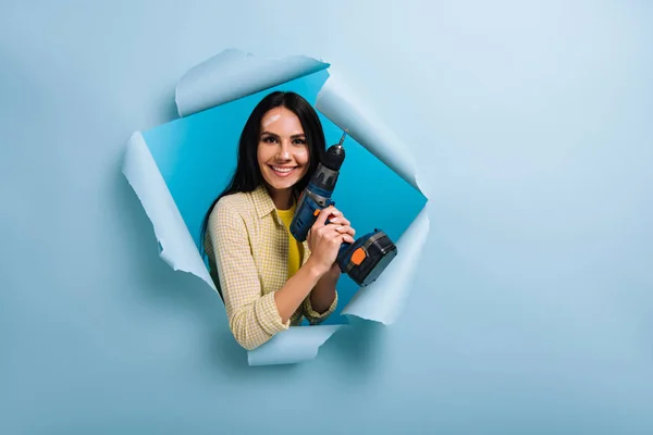 Handywoman alegre com rosto sujo em pintura segurando broca elétrica em papel rasgado, isolado em azul — Fotografia de Stock