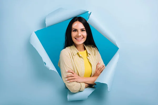 Confident workwoman with crossed arms and dirty face in paint standing in torn paper, isolated on blue — Stock Photo