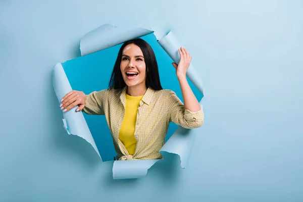 Happy workwoman with dirty face in paint standing in torn paper, isolated on blue — Stock Photo
