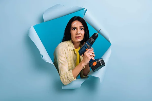 Scared female manual worker with dirty face in paint holding electric drill in torn paper, isolated on blue — Stock Photo